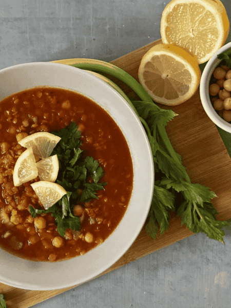 Egy tál gazdag, sűrű marokkói paradicsomleves csicseriborsóval és lencsével, friss petrezselyemmel, citromkarikákkal egy vágódeszkán tálalva.